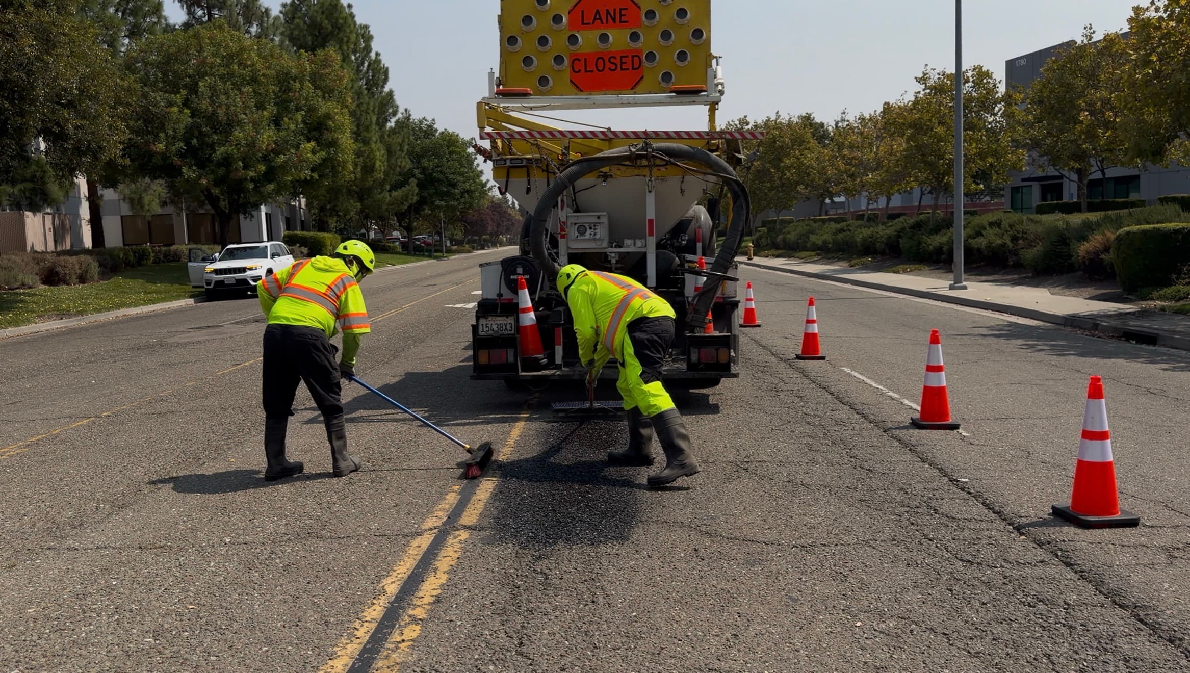 Two operators mid-repair: one injecting, one brooming, with the machine and lane-closed sign visible